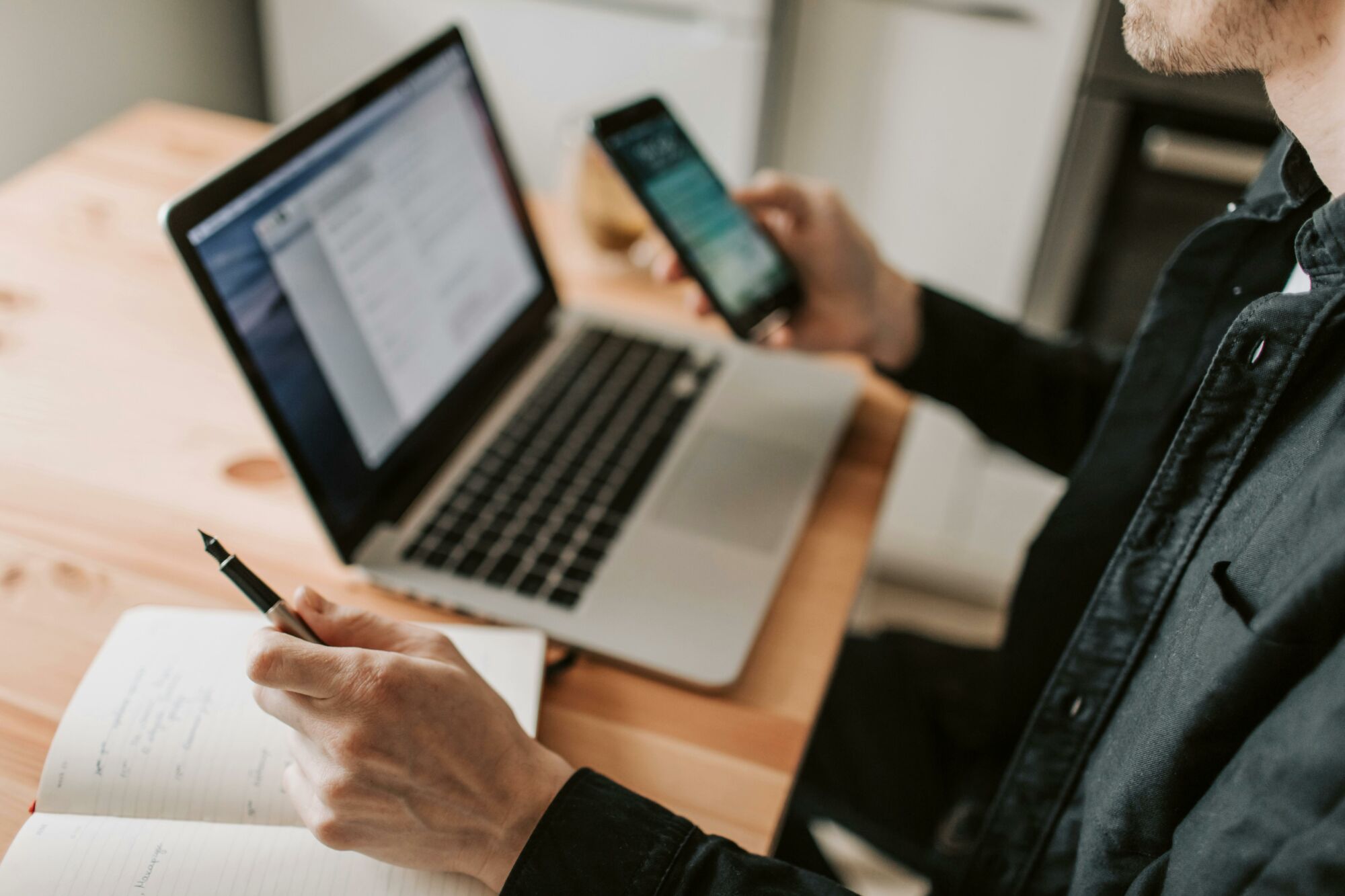 A person multitasking with a laptop and smartphone at a wooden desk.